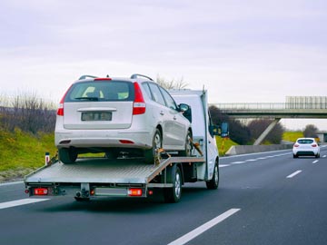 Ein weißes Auto wird auf einem Abschleppwagen auf der Autobahn transportiert, während eine Brücke im Hintergrund unter einem bewölkten Himmel sichtbar ist.