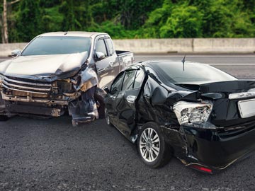 Ein silberner Pickup und ein schwarzer Sedan sind in einen schweren Unfall verwickelt, wobei beide Fahrzeuge auf der Straße erhebliche Schäden aufweisen.